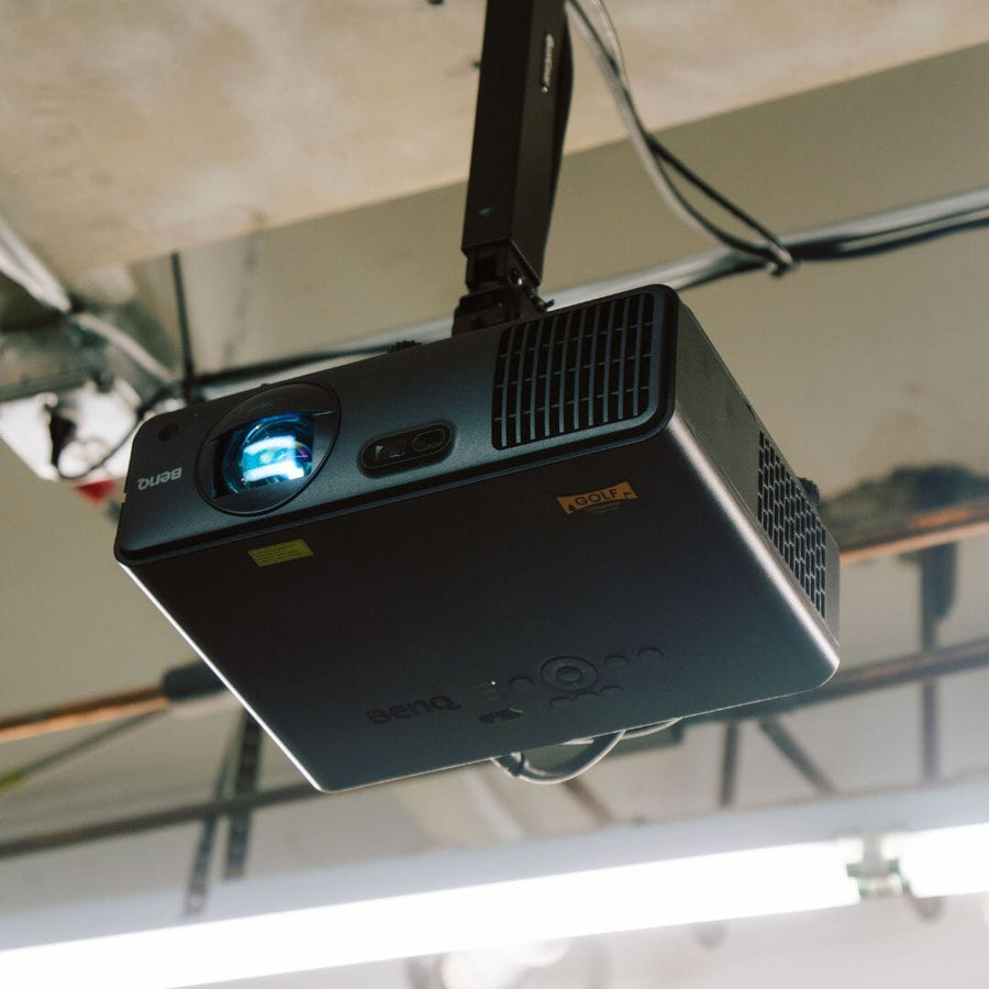 BenQ AH700ST golf simulator projector mounted to a ceiling, with its lens glowing blue